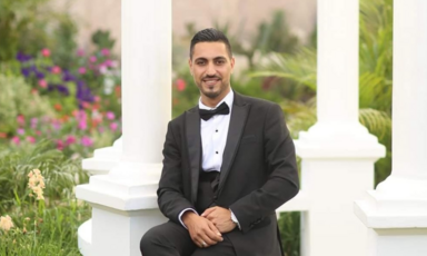 A man wearing a tuxedo poses, sitting and smiling, in a garden.