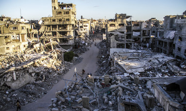 Landscape view of people walking and biking on narrow road cleared of debris between destroyed and bombed-out multi-story buildings