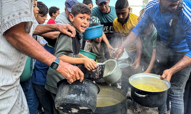 A boy looks at the camera while standing close to other boys and men holding pots over a vat of soup