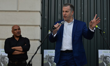 A man wearing a dark blue suit and a white shirt holds a microphone as another man stands behind him 