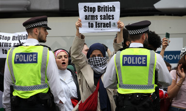 A protester holds up a sign saying Stop British Arms Sales to Israel as she stands behind police and beside other protesters 