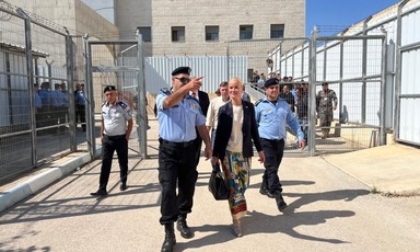 A woman stands between two men in uniform with a large building and a large gate partly topped with barbed wire behind them