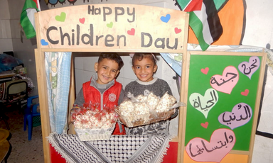 A boy and girl hold baskets while standing behind frame decorated with Palestinian symbols