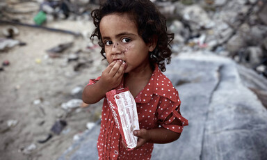 A young girl holds a small bag in one hand while raising another hand to her mouth