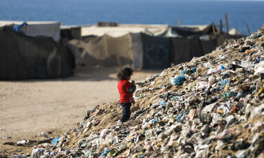 A young girl stands on top of a garbage pile with makeshift shelters and the sea in the background