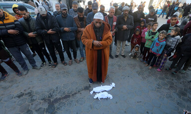 A man wearing a dark orange coat prays over two tiny bodies in white shrouds. A crowd of mourners is behind him.