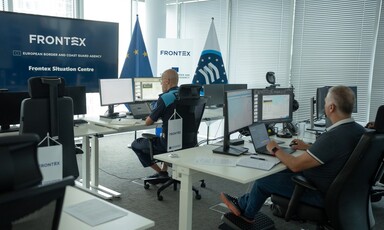 Two men sit behind computer screens and a banner reading Frontex European Border and Coast Guard Agency