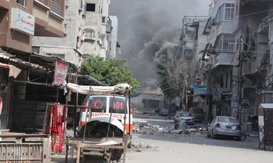 An ambulance in foreground and huge dust cloud in background caused by Israeli bombing