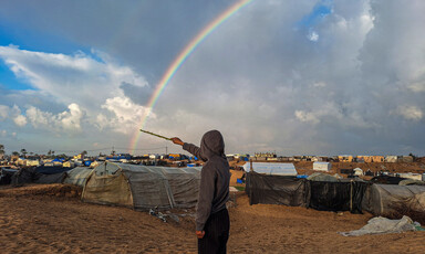 Amidst tents of displaced families, a boy points a stick at a rainbow arching across the sky. 