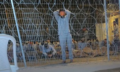 Men blindfolded and bound sit or stand behind razor wire fence