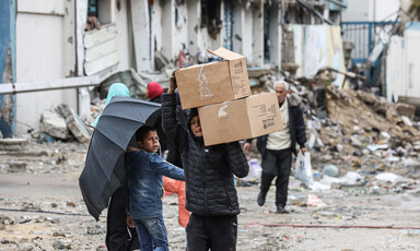 A boy holding an umbrella looks at another boy who is carrying boxes on his shoulders