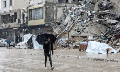 A Palestinian youth, wearing black jeans and a black sweatshirt, carries an umbrella past piles of rubble