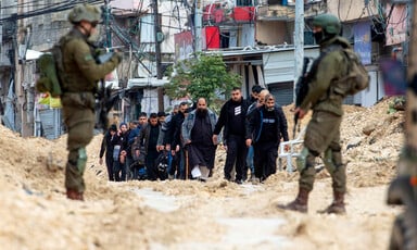 People march on a dirt road as they're watched over by two Israeli soldiers