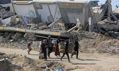 Landscape view of a family of six carrying rolled-up mattresses and buckets while walking on road lined by destroyed buildings