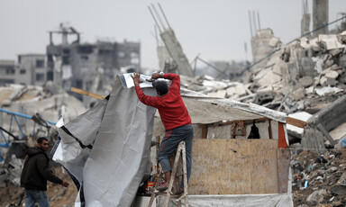 A man nails a tarp cover onto a wooden structure surrounded by rubble