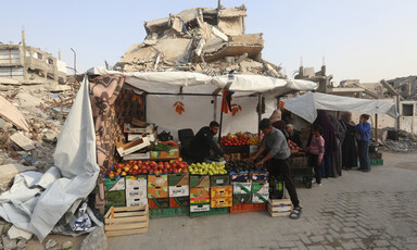 Palestinians shop at a small produce stall in a canvas tent in front of destroyed buildings