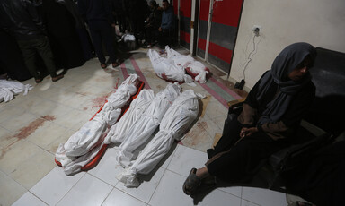A woman sits on the floor leaning against a wall next to the shrouded bodies of several children