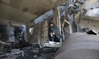 Inside a gutted-out concrete building stands a woman, a plume of smoke and dust rising to her left. 