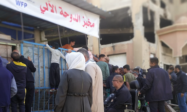 A sign for the Doha Bakery, in Arabic, and a crowd of people gather around it, and children climb on gates
