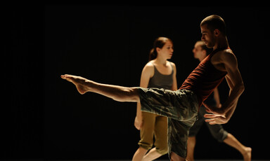 A man lifts his foot as part of a dance routine in front of two other people on a stage 