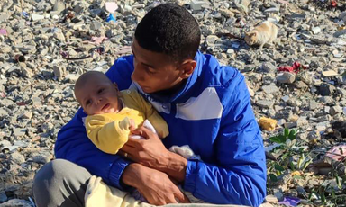 A man in a blue jacket cradles a newborn baby in a yellow jumper amid piles of rubble