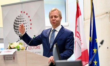 Diplomat wearing a suit stands at a lectern with two flags behind him 
