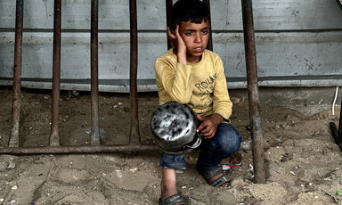 A boy in a yellow shirt kneels on the ground with an empty pot in his hands