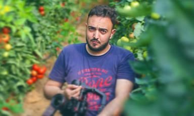 Young man wearing t-shirt holds camera while standing next to tomato plants