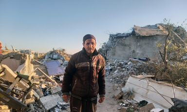 An older man looks at the camera while standing on narrow road in between destroyed buildings