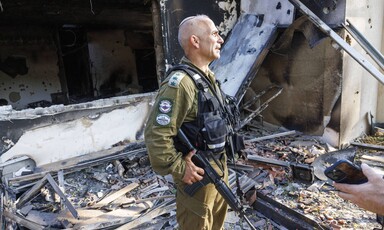 Man in military uniform stands in front of badly burned and damaged house