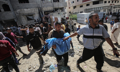 A group of men walk together, one of them is carrying a body in a blue cloth