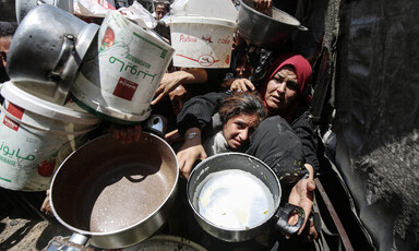 Girl looks towards camera while in the arms of woman holding an empty pot in crowd