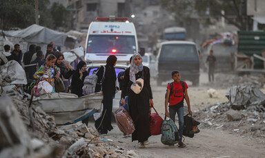 Women and children carry belongings amidst rubble. Ambulances are behind them.