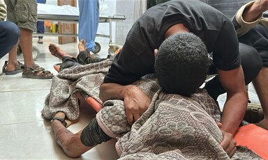 A man cradles the body of a loved one on the floor of a hospital. The body is wrapped in a blanket.