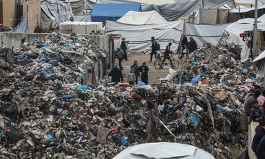 Piles of trash make people walking by look tiny, tents in the background