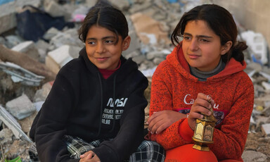 Two young girls sit on rubble, one holding a small lantern