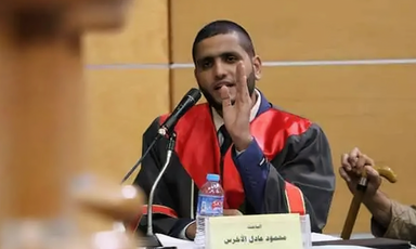 A man wearing graduation regalia speaks into the microphone. 