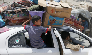 A boy sits on a car's window while holding boxes strapped to the top of the vehicle