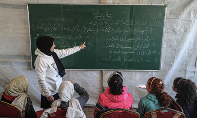 A teacher in a tent points to a blackboard as she stands in front of several children 