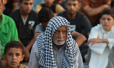 A close-up of an elderly man with a black-and-white checkered kuffiyeh draped over his head. 