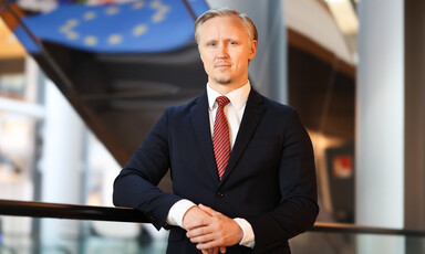 A man wearing a suit and tie stands in front of the blue and yellow logo of the European Union