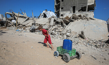 A child, dressed in red, pulls a can of water on a small cart up a hill, surrounded by rubble of destroyed buildings.