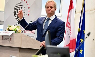 A man wearing a suit stands at a podium with flags behind him 