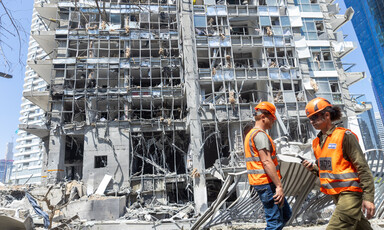 Two workers stand in front of badly damage facade of tall building