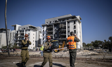 Personnel in uniform stand in front of damaged apartment buildings