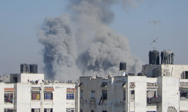 In daytime, massive clouds of black-gray smoke appear over city buildings