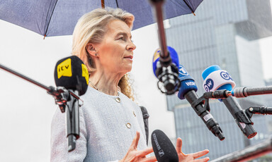 European Commission President Ursula von der Leyen stands beneath an umbrella and surrounded by television microphones 