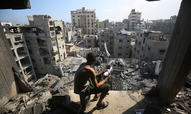 A young man holding a notebook sits on a chair overlooking a cityscape of destroyed buildings