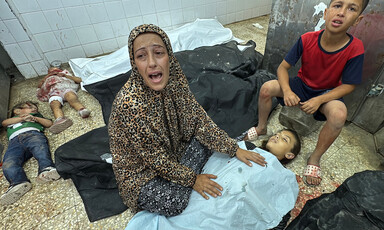 A woman wearing prayer clothes mourns over the body of a small child under a blue medical blanket. Two small children lie motionless behind her as an older boy sits next to her.
