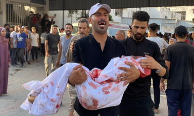 A man in a black t-shirt carries the body of a child, wrapped in a bloodied white shroud, as other people in the background look on.